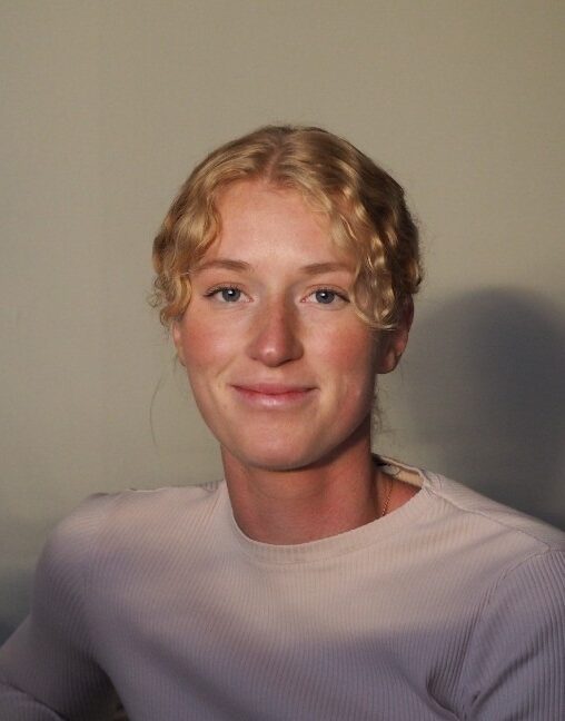 En person med lockigt blont hår och ljusrosa tröja ler medan hen tittar mot kameran, sittande framför en enkel bakgrund.