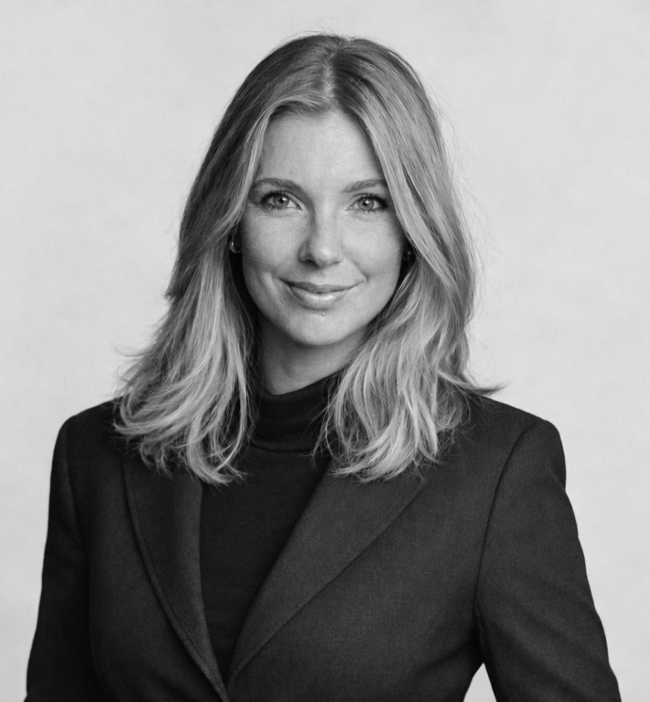 En kvinna med axellångt ljust hår i mörk kavaj och polotröja poserar för ett svartvitt porträtt mot en enkel bakgrund.