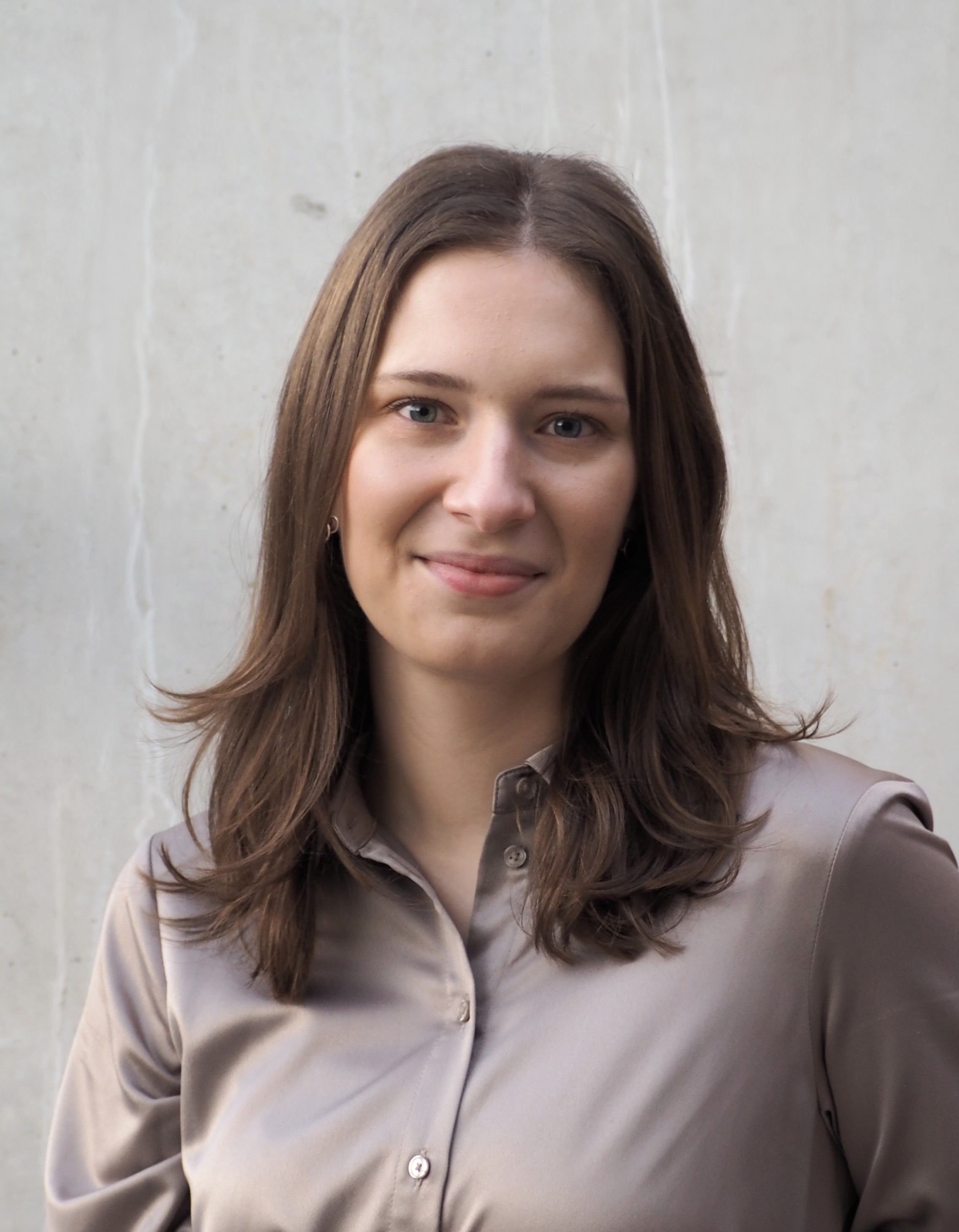 En kvinna med rakt brunt hår och taupe skjorta står framför en ljusgrå betongvägg och ser mot kameran med ett litet leende.