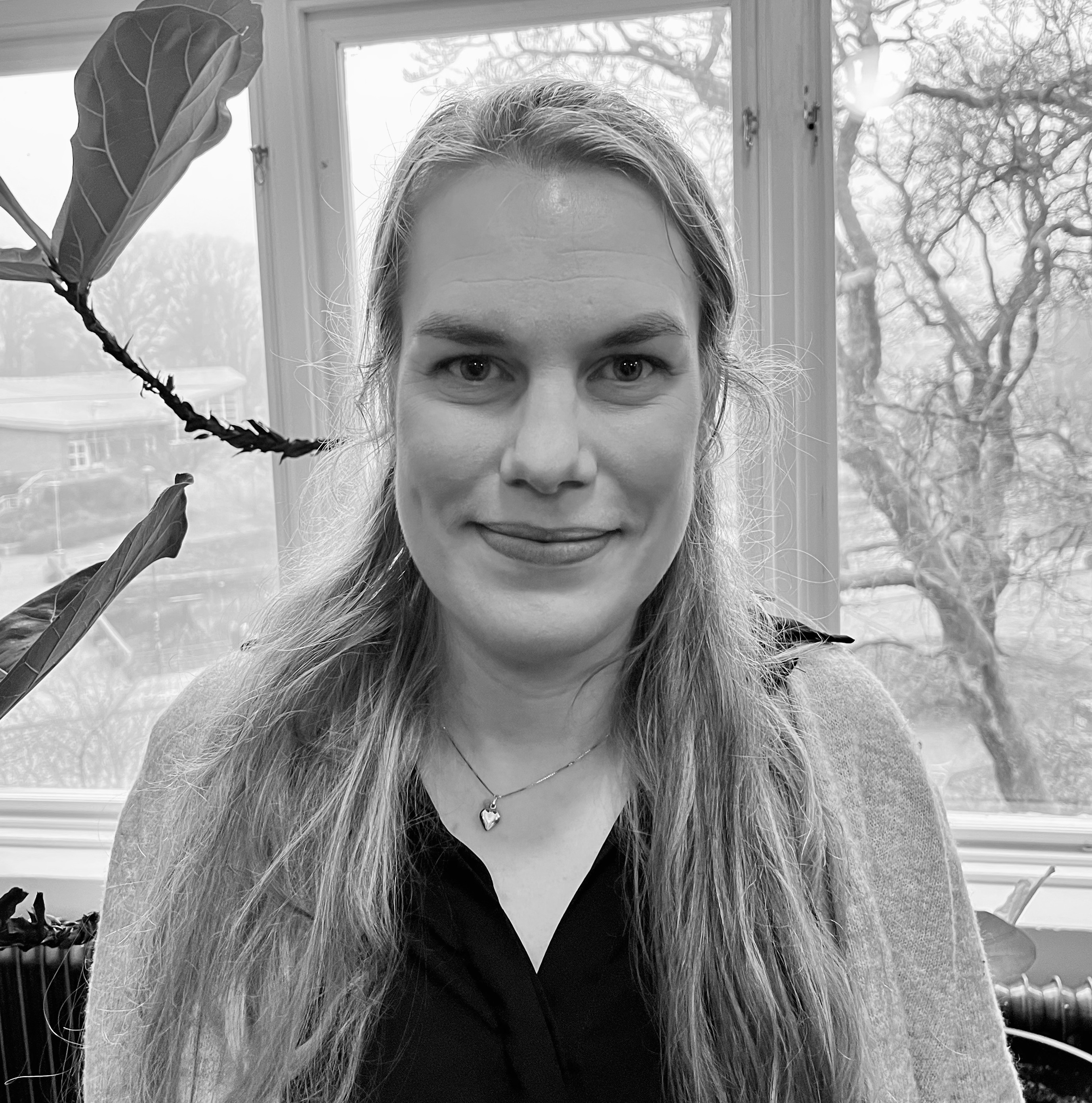 En kvinna med långt hår står inomhus framför ett fönster, har på sig ett halsband och en mörk tröja, träd och byggnader syns utanför.