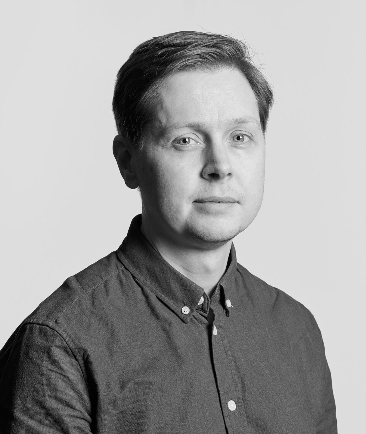 En man med kort hår och skjorta tittar mot kameran med neutral min. Bakgrunden är enfärgad.