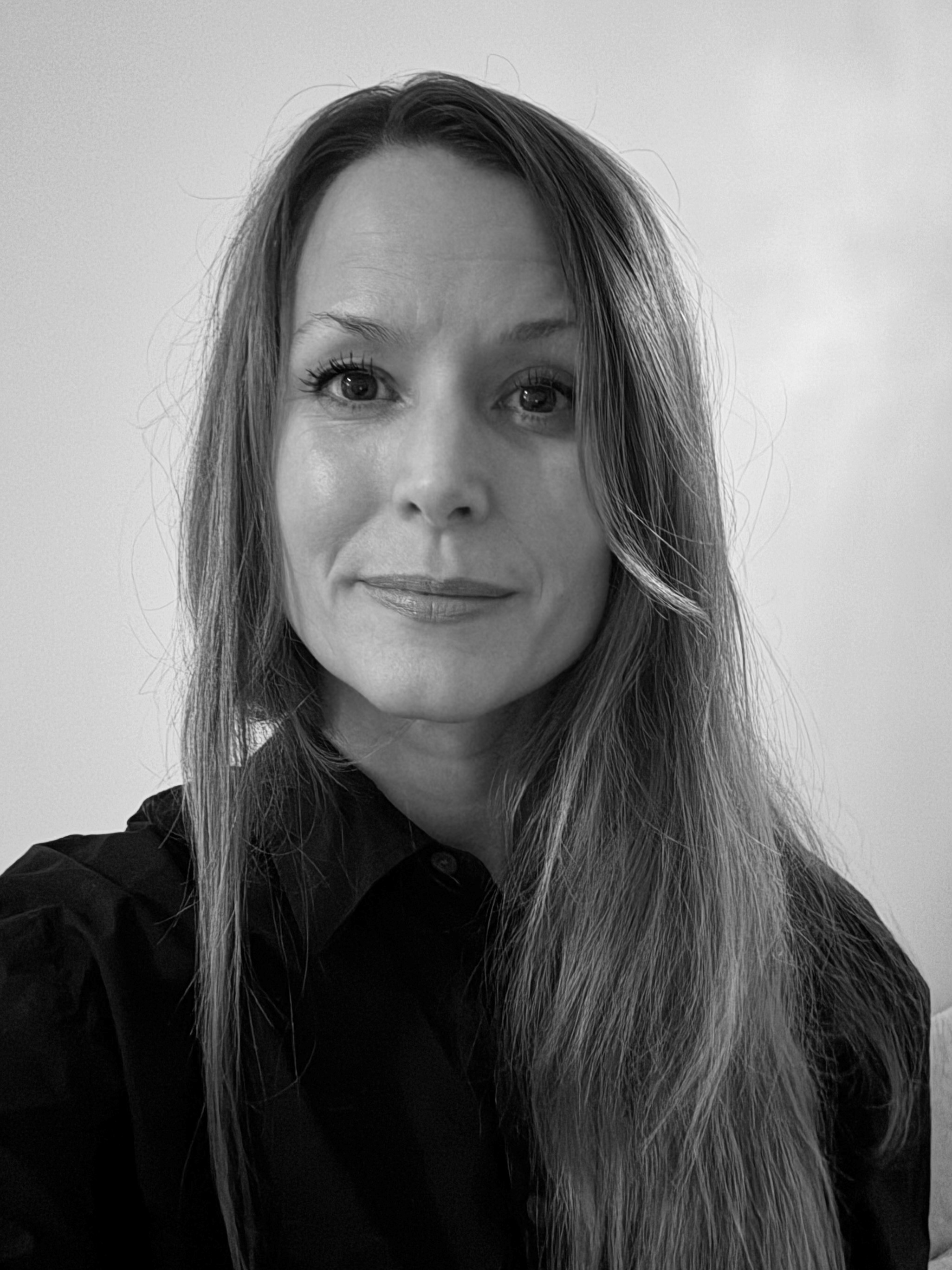 En kvinna med långt rakt hår och mörk skjorta tittar mot kameran med neutral min. Bilden är i svartvitt.