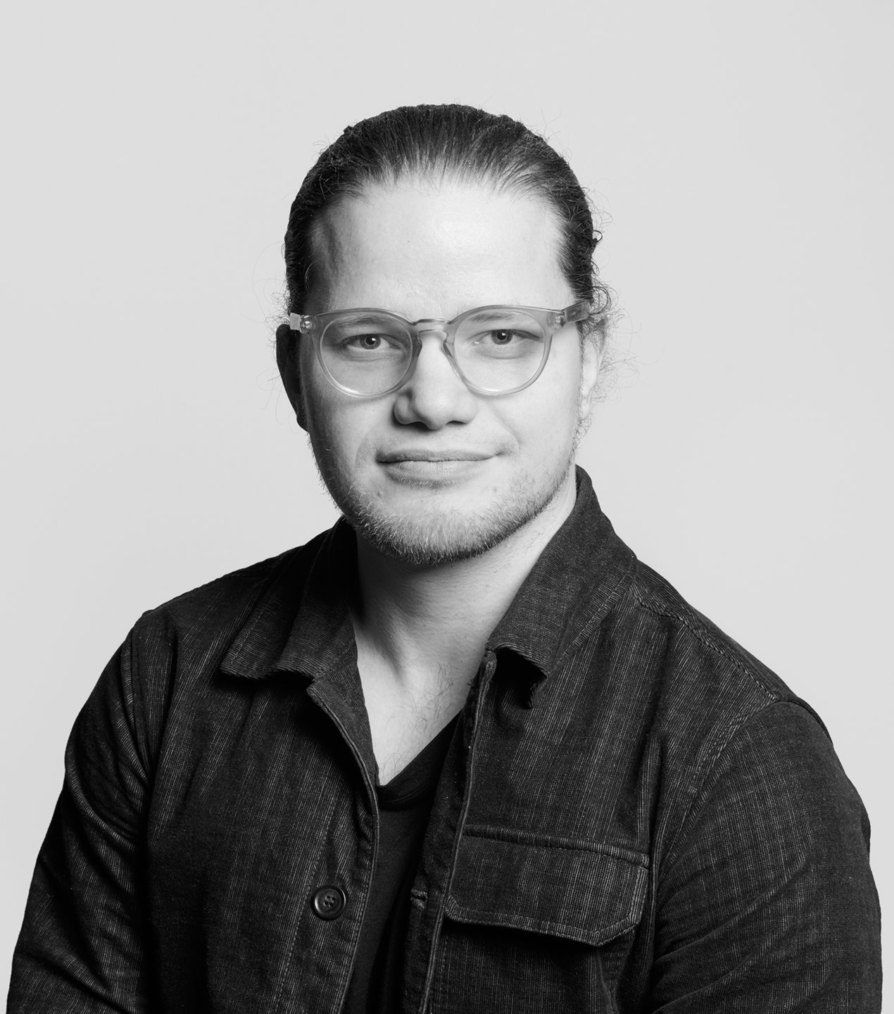 En person med glasögon och långt bakåtsamlat hår har en mörk jacka, tittar mot kameran med neutral min mot ljus bakgrund.