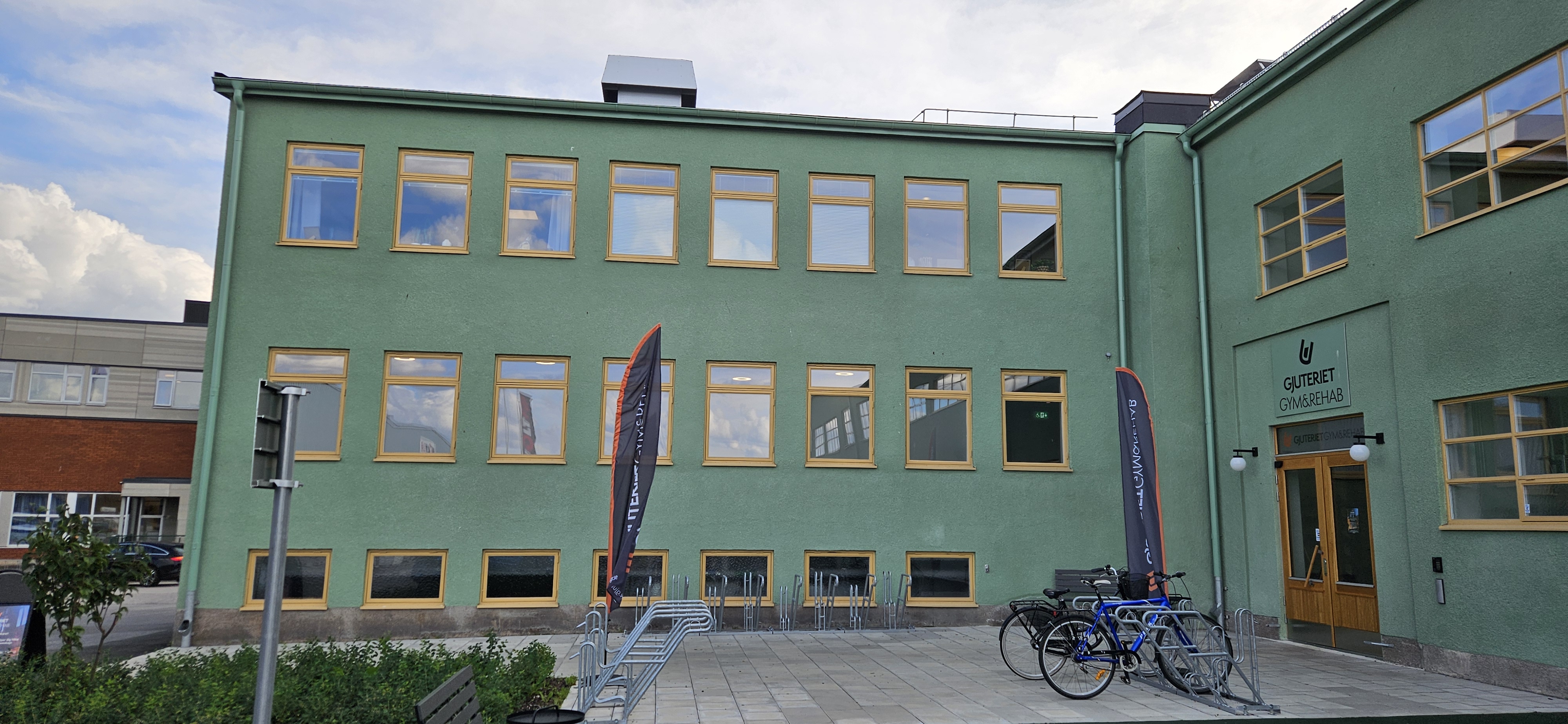 En trevåningsbyggnad med fönster med gula ramar, två cyklar vid ingången och två vertikala banderoller nära dörren.