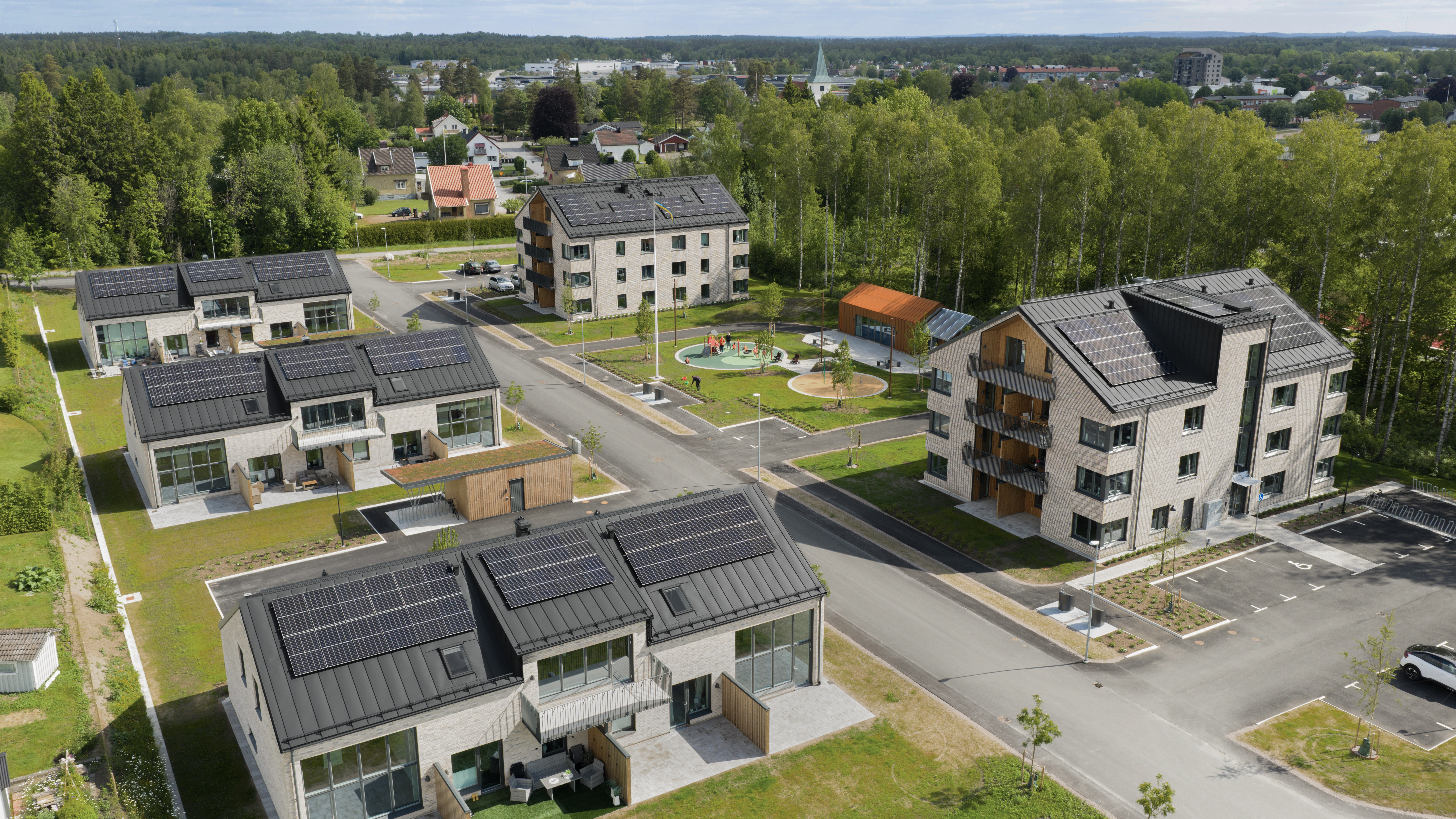 Flygfoto över ett bostadsområde med hus, lägenhetsbyggnader, solpaneler på taken och en lekplats omgiven av träd.