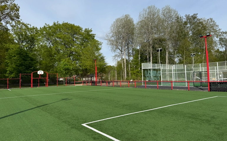 Utomhusplan med konstgräs, röd inhägnad, basketkorg och tennisbanor i bakgrunden, omgiven av träd under klar himmel.