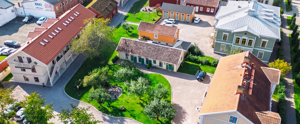 Flygbild över ett bostadsområde med flera hus, gräsmattor, träd, parkerade bilar och asfalterade gångvägar.