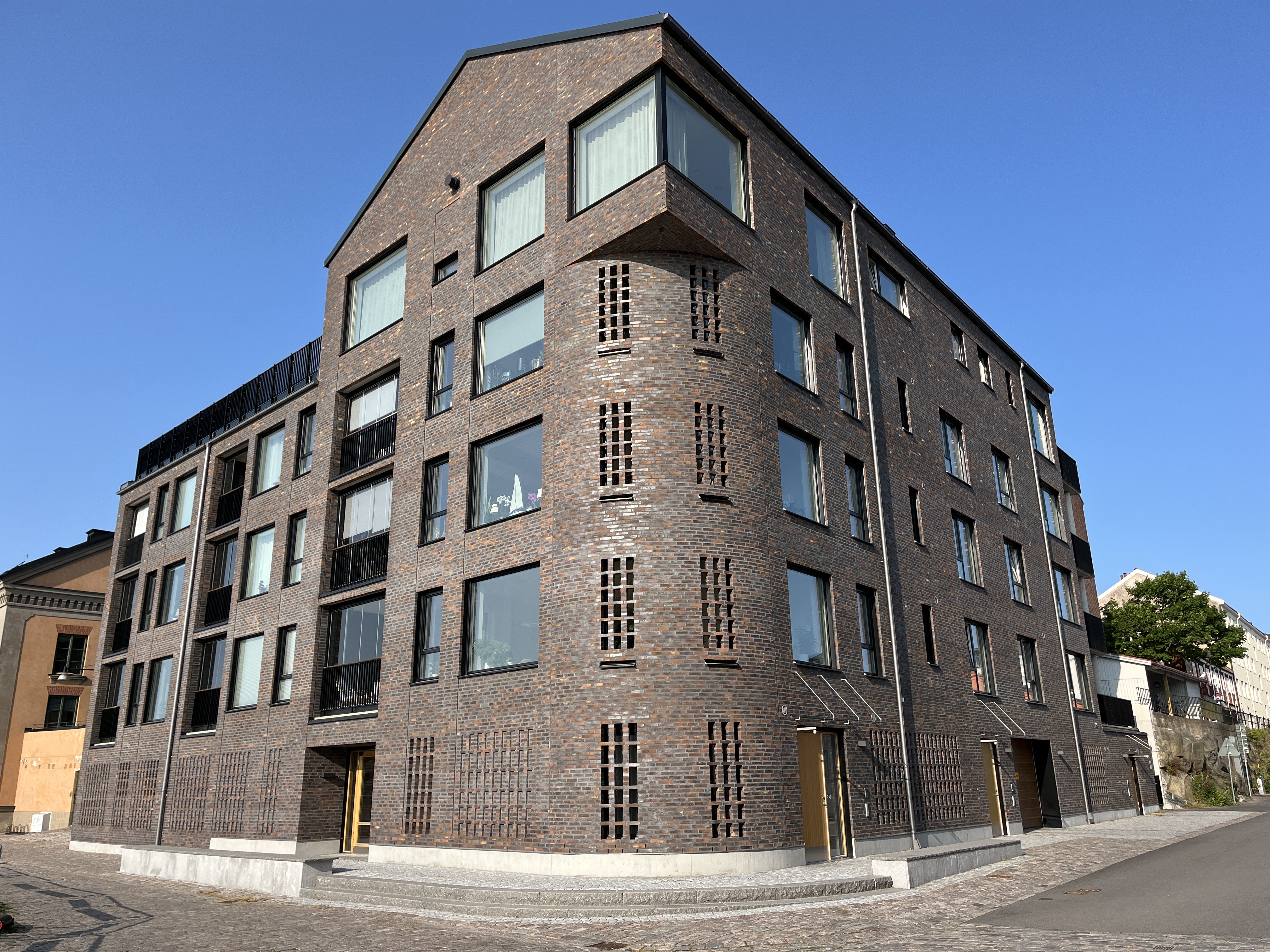 Ett fyravånings hyreshus i tegel med stora fönster och geometriska former, placerat i ett hörn vid en kullerstensgata.