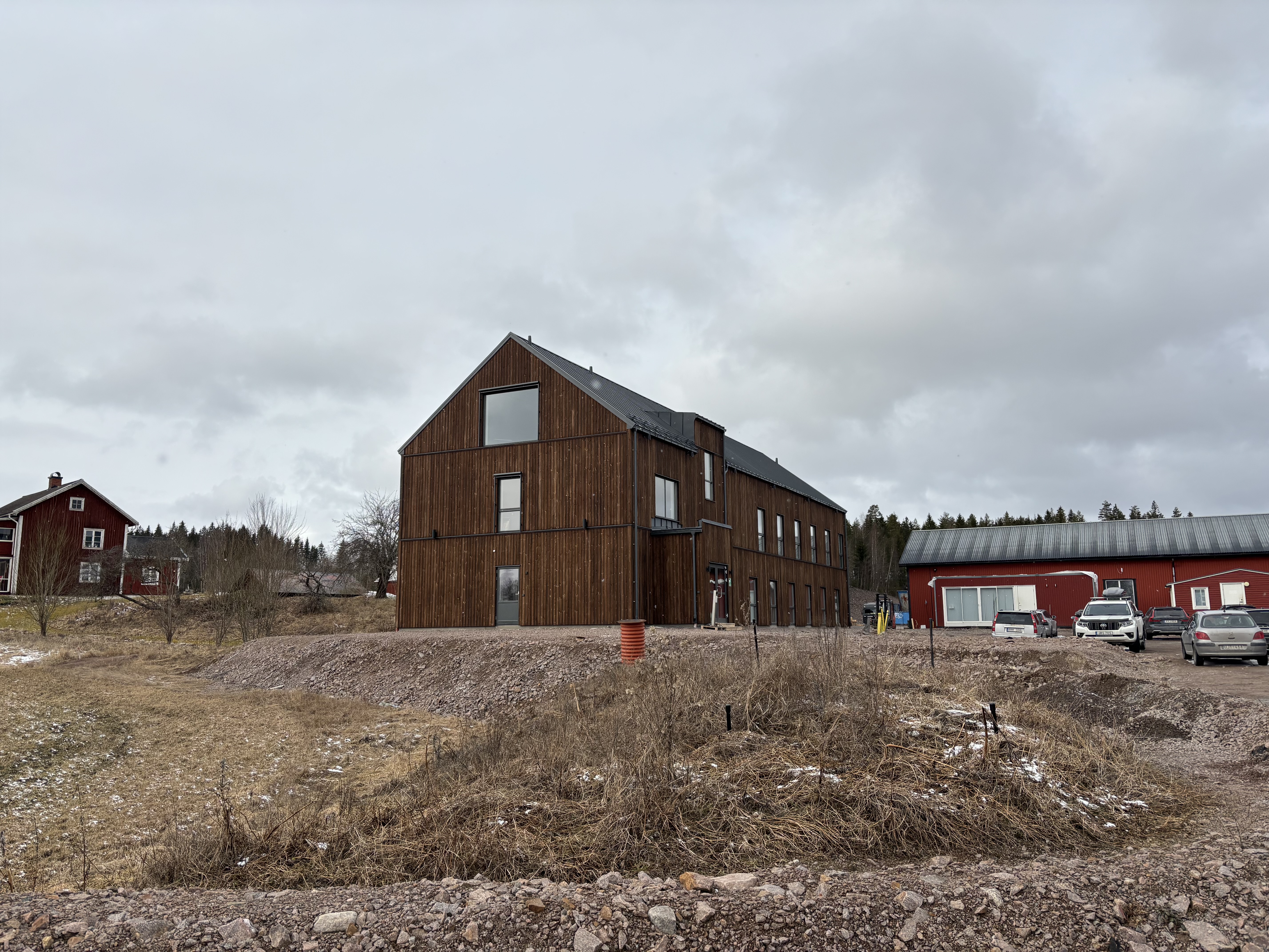 En byggnad i trä med stora fönster står på en upphöjd tomt vid en grusplan, med röda lador och bilar i bakgrunden.