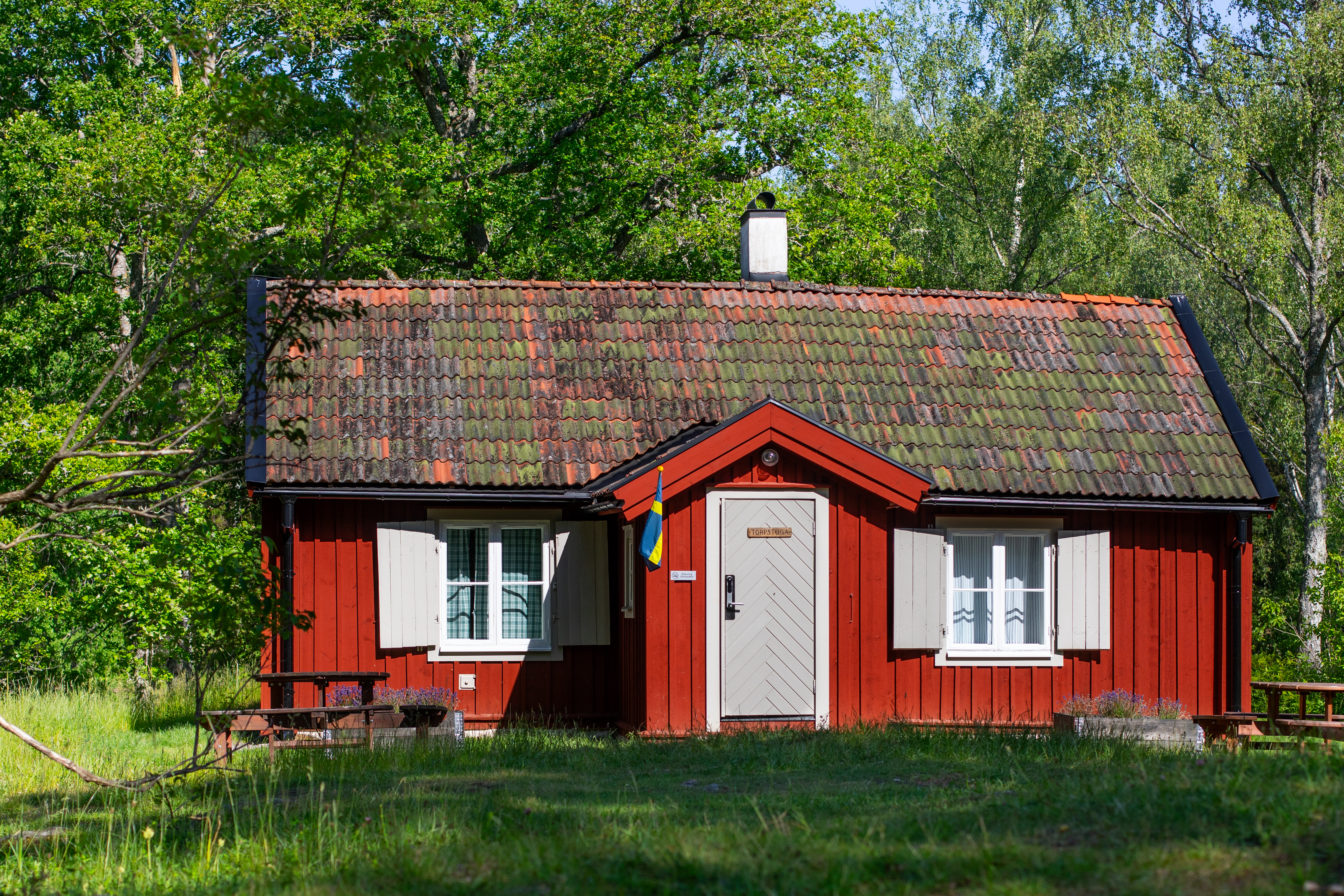 Ett rött trähus med vita kanter, tegeltak och en svensk flagga står på gräs, omgiven av träd.