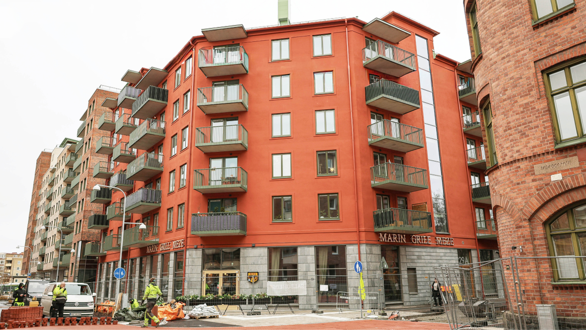 Ett rött flerfamiljshus med flera balkonger bredvid en tegelbyggnad; byggarbetare och utrustning syns i förgrunden.