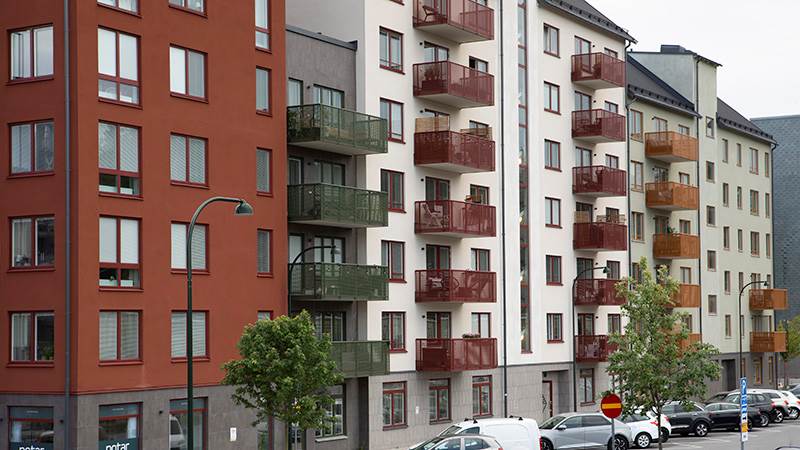 Ett lägenhetshus med balkonger och bilar parkerade längs gatan. Träd står vid trottoaren en molnig dag.