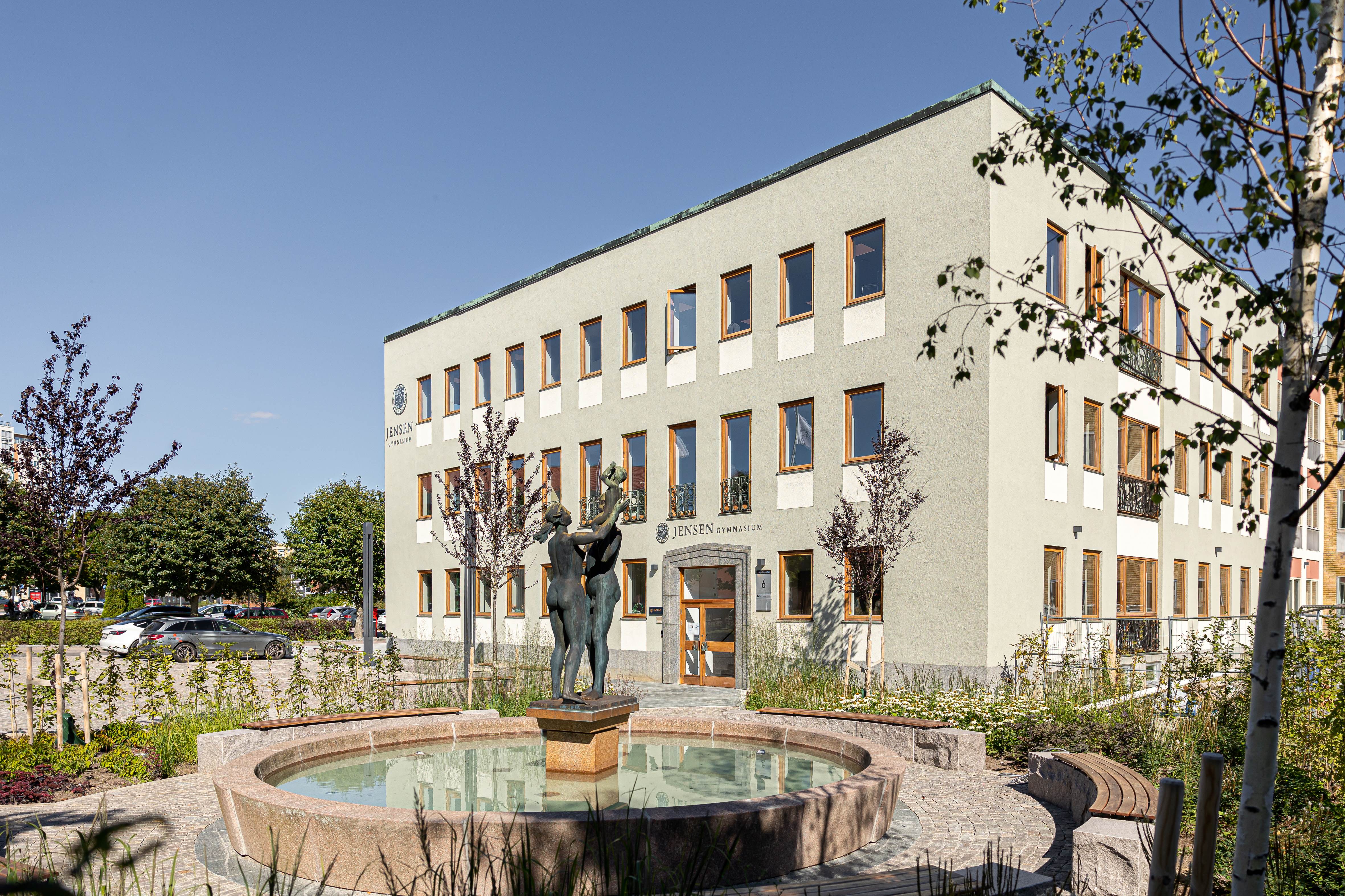 En byggnad i tre våningar med många fönster, en huvudentré och en rund fontän med staty framför, omgiven av träd och parkering.