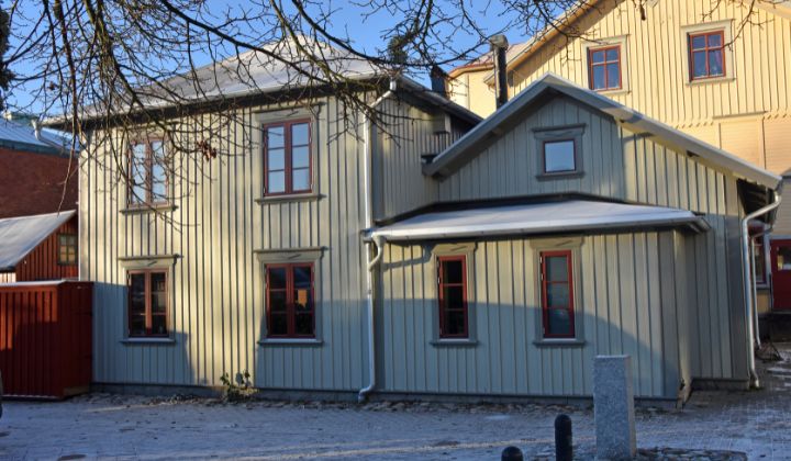 Ett tvåvånings trähus i ljusgul färg med rödramade fönster, snedtak och synliga stuprör, fotograferat en klar dag.