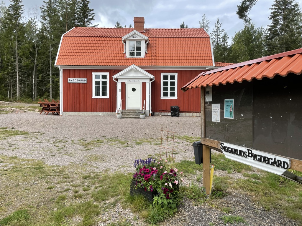 En röd träbyggnad med vit dörr och fönsterfoder märkt "BYGDEGÅRD" står på landsbygden med träd, anslagstavla, blommor och picknickbord utanför.