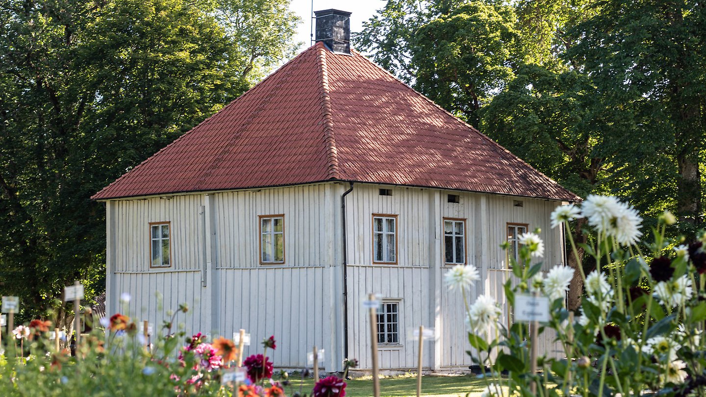 Ett vitt trähus med rött tegeltak står bland träd och blommor i en trädgård under en solig dag.