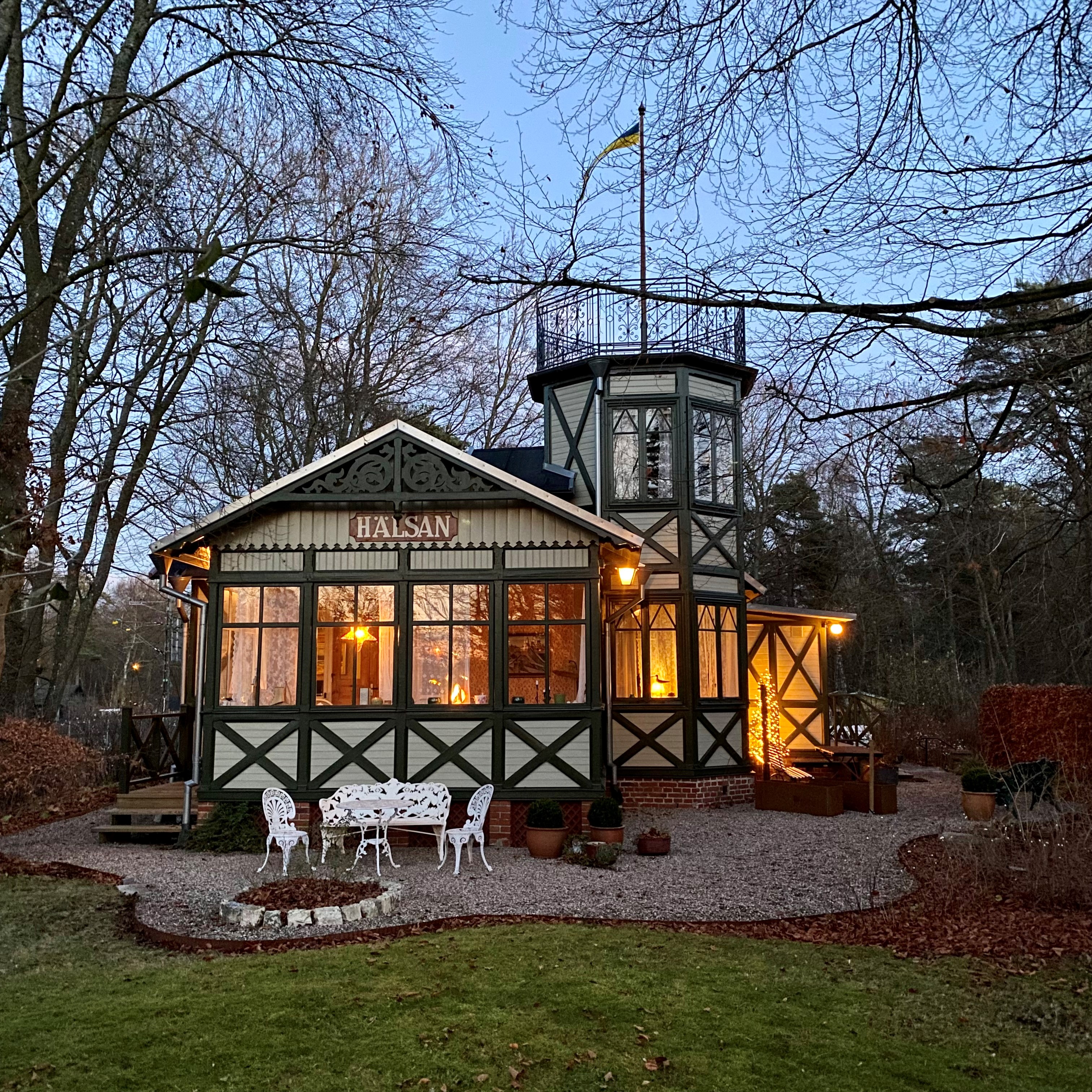 Ett tvåvånings trähus med lampor tända, omgiven av träd och grusgård, vita utemöbler framför och svensk flagga på taket.