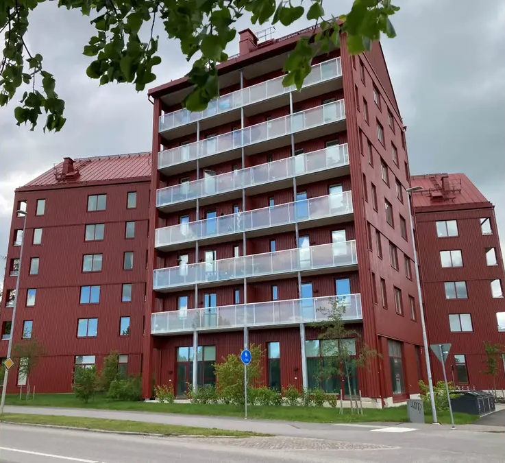 Ett rött flerfamiljshus med flera balkonger och stora fönster, omgiven av växter och en asfalterad väg framför.