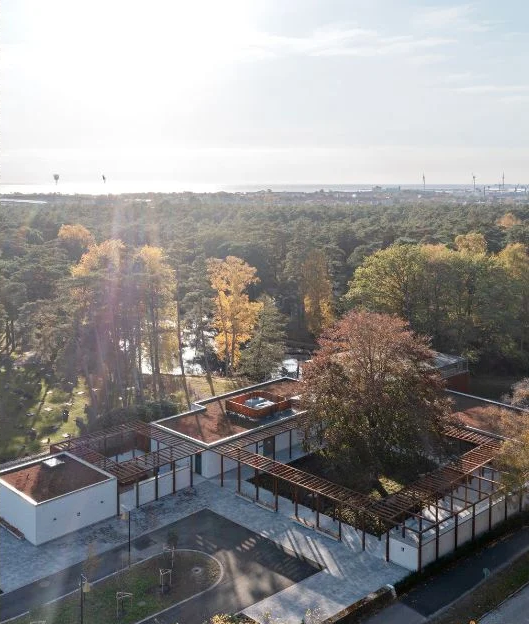 En modern byggnad med platta tak och pergolor omges av träd med höstlöv, med skog och vatten i bakgrunden under en ljus himmel.