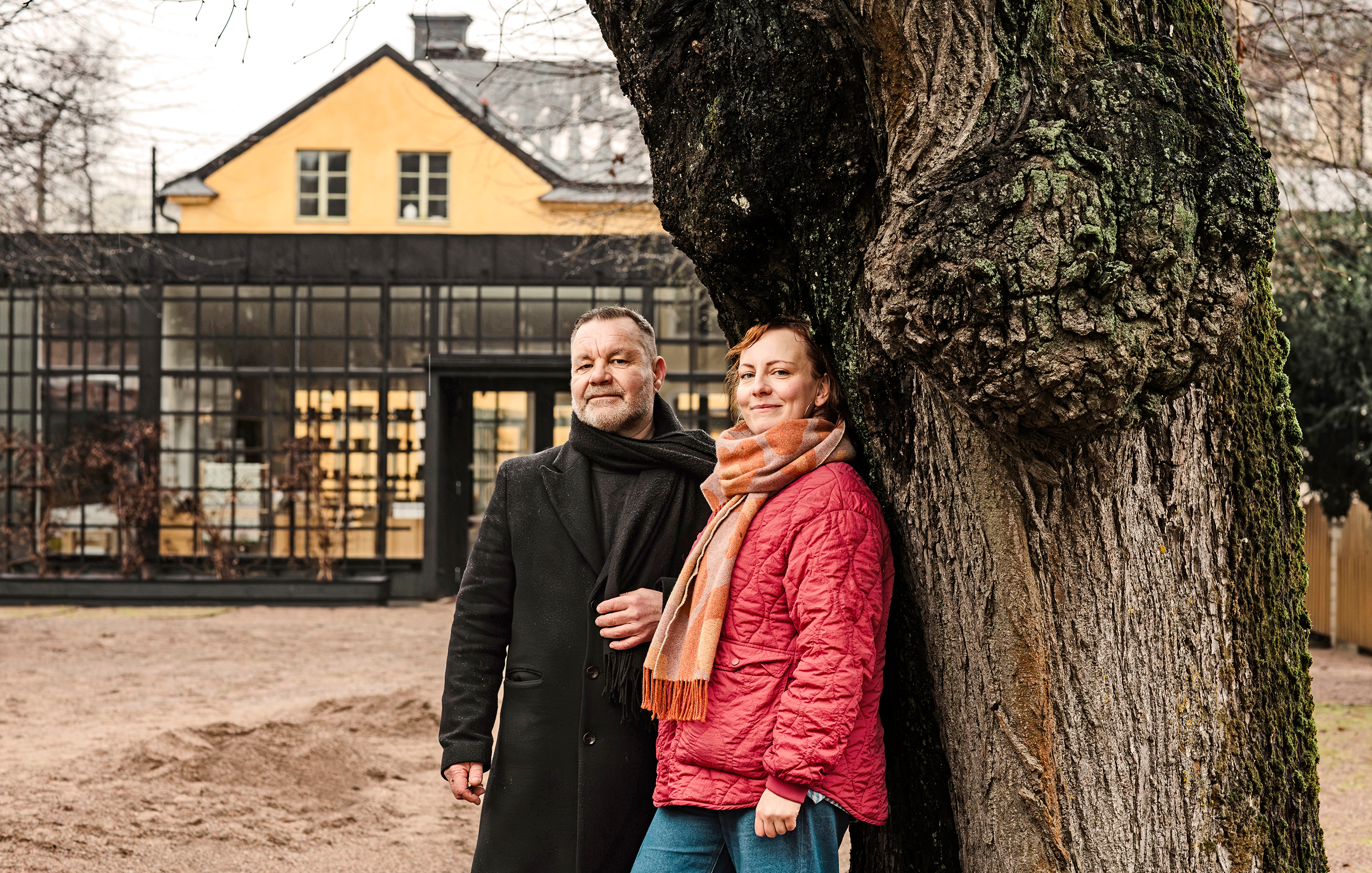 En man och en kvinna står bredvid ett träd utomhus, med en gul byggnad och ett växthus i glas i bakgrunden.