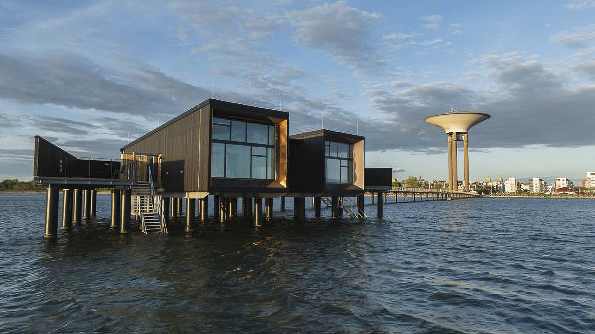 Byggnader på pålar står över vatten nära stranden, med ett stort vattentorn och stad i bakgrunden under delvis molnig himmel.