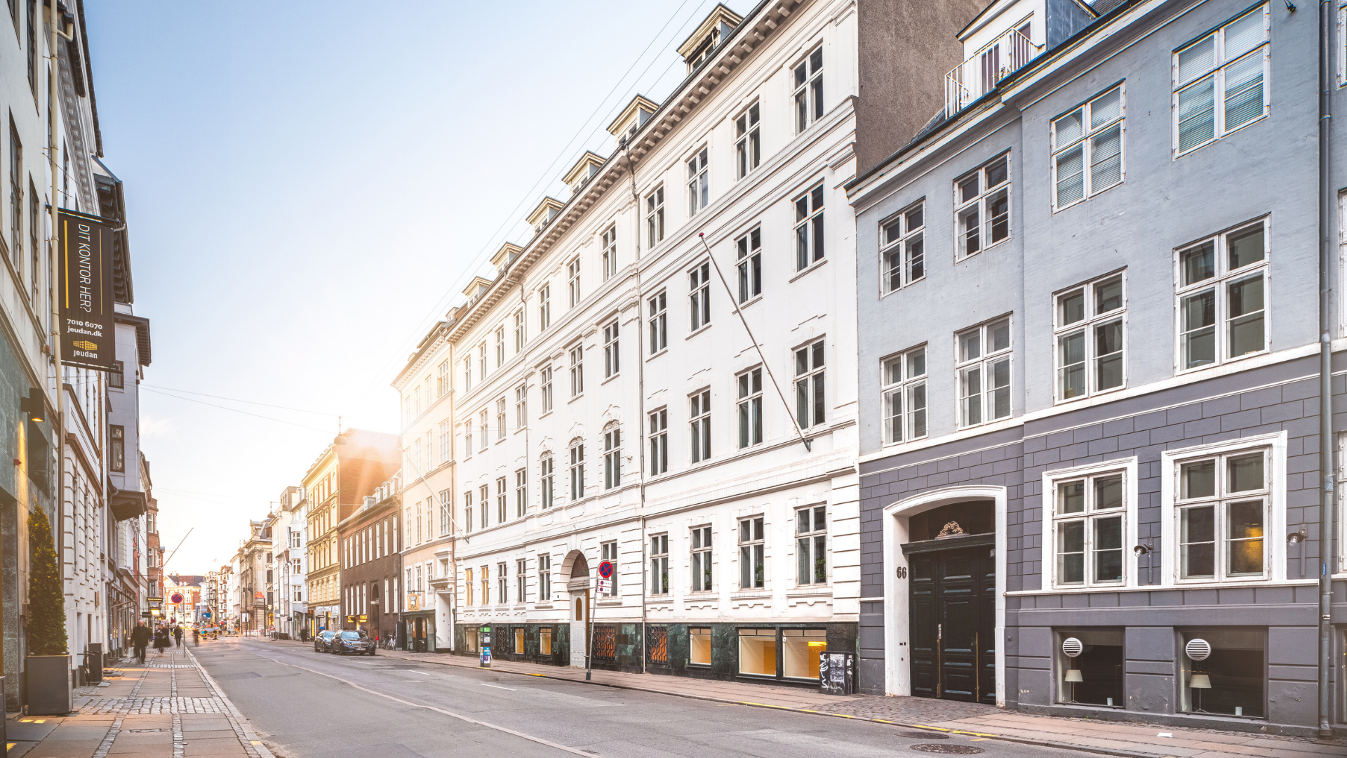 En stadsgata med flera höghus i flera våningar, stora fönster och ljusa fasader under en klar himmel.