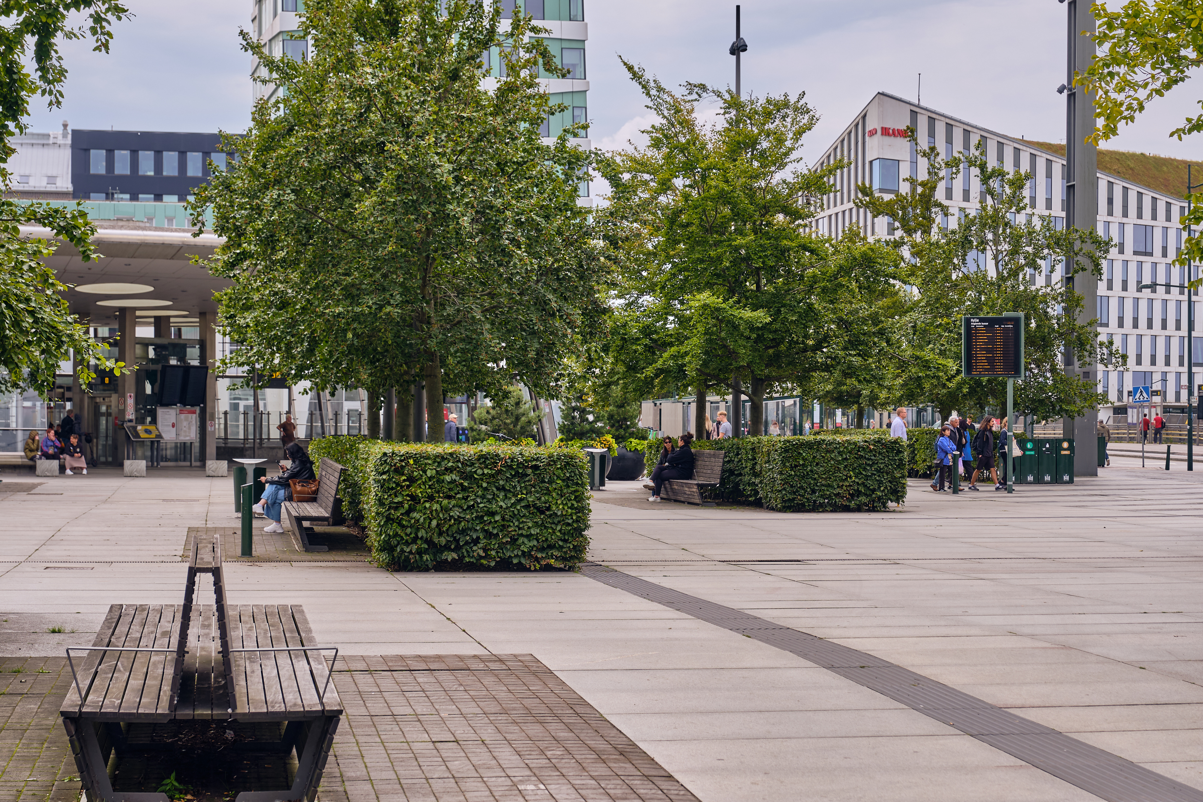 Stadstorg med bänkar, träd och häckar; personer sitter och går; byggnader syns i bakgrunden.