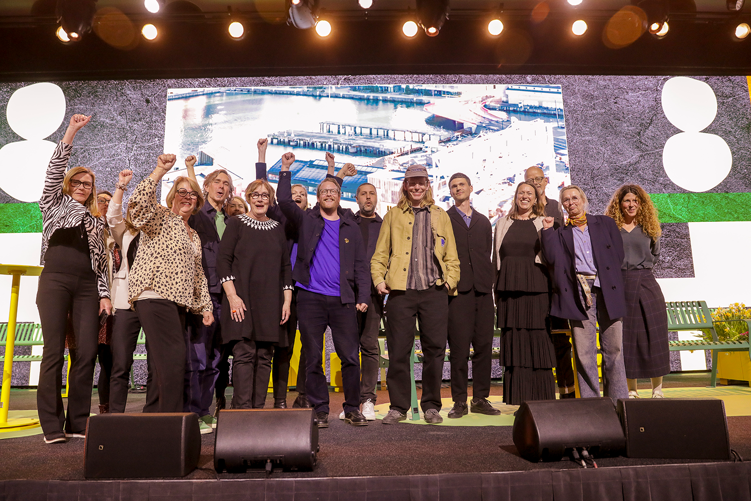 En grupp personer står på en scen, några lyfter sina nävar, framför en stor skärm med ett flygfoto och abstrakta mönster.