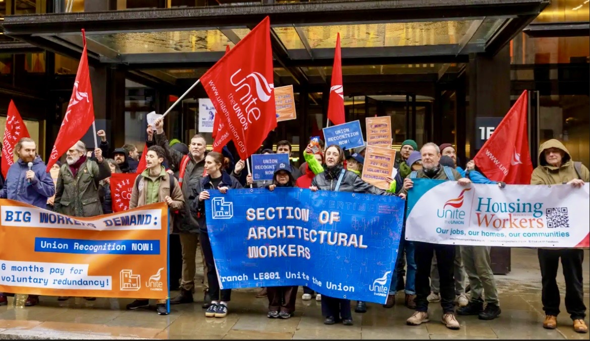 En grupp personer står utomhus med banderoller och flaggor för fack inom arkitektur och bostäder under en protest eller strejk.