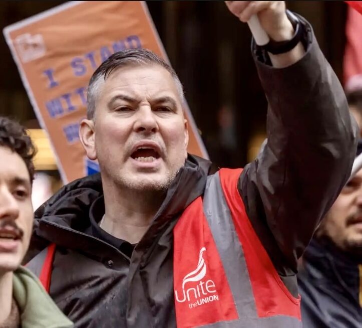 En man med en röd "Unite the Union"-väst ropar ropar på en protest och lyfter armen; en skylt med texten "I stand with" syns bakom.
