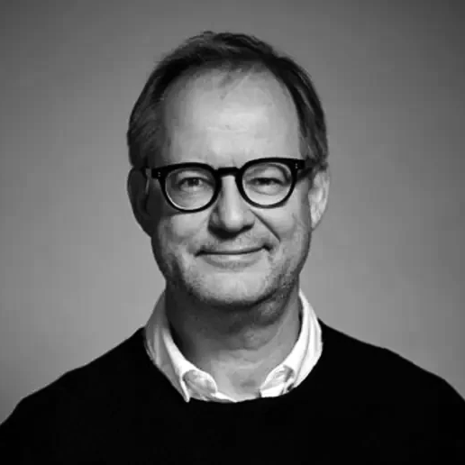 En medelålders man med kort hår och glasögon, iförd skjorta och mörk tröja, står mot en enfärgad bakgrund.