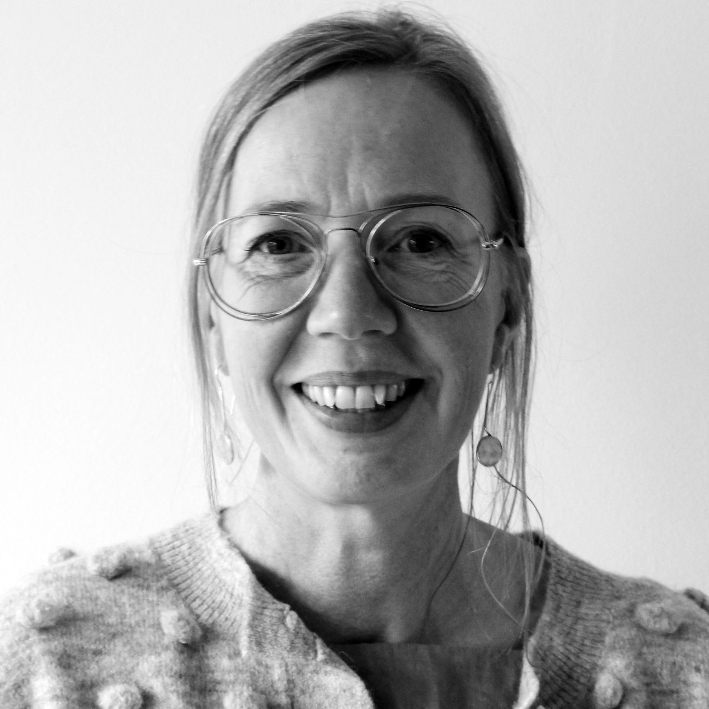 Porträtt i svartvitt av en kvinna med glasögon, ljus tröja, långa örhängen och långt bakåtdraget hår som ler mot kameran.