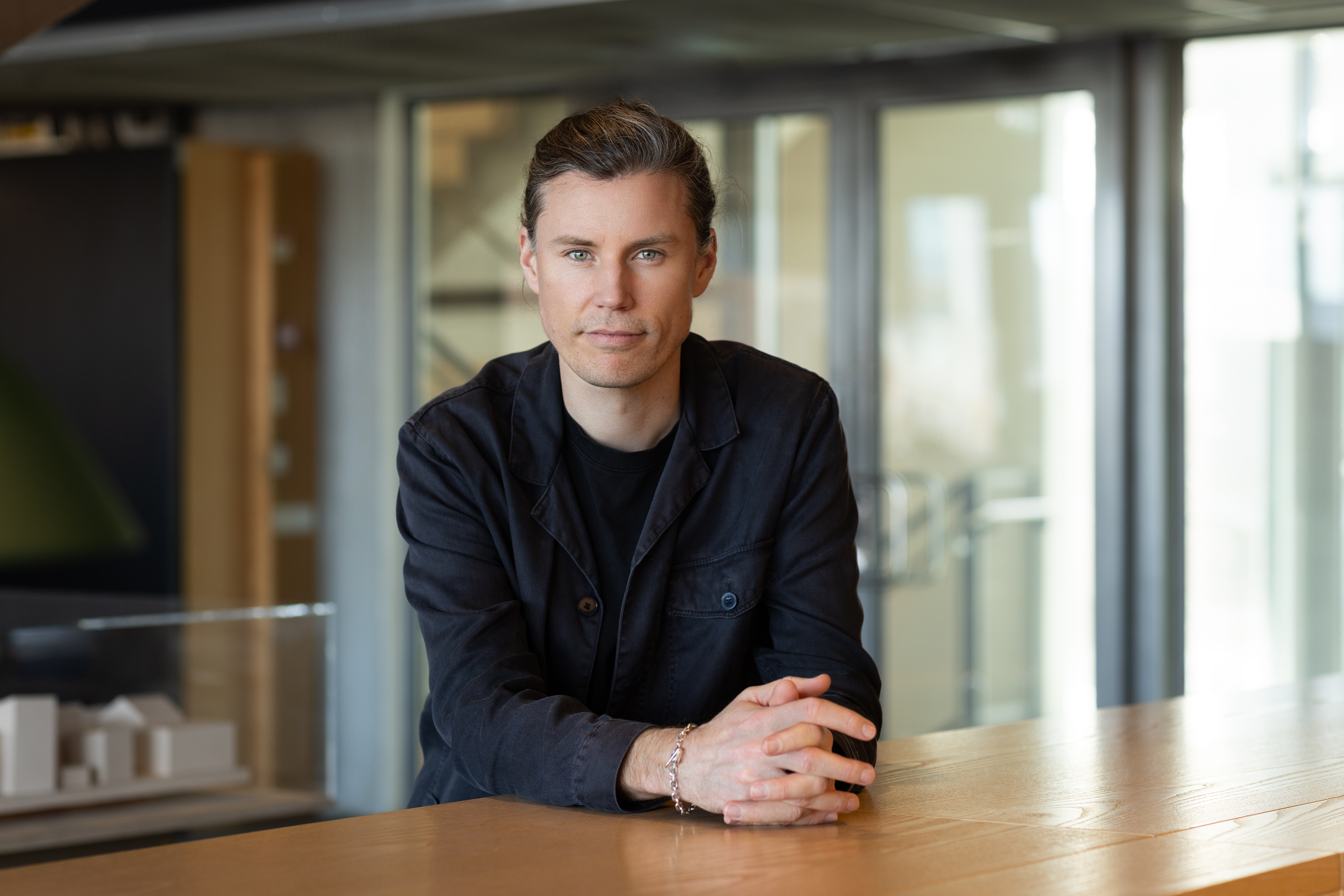 En person med ljus hud och brunt hår sitter vid ett träbord, har en mörk jacka på sig och tittar mot kameran inomhus.