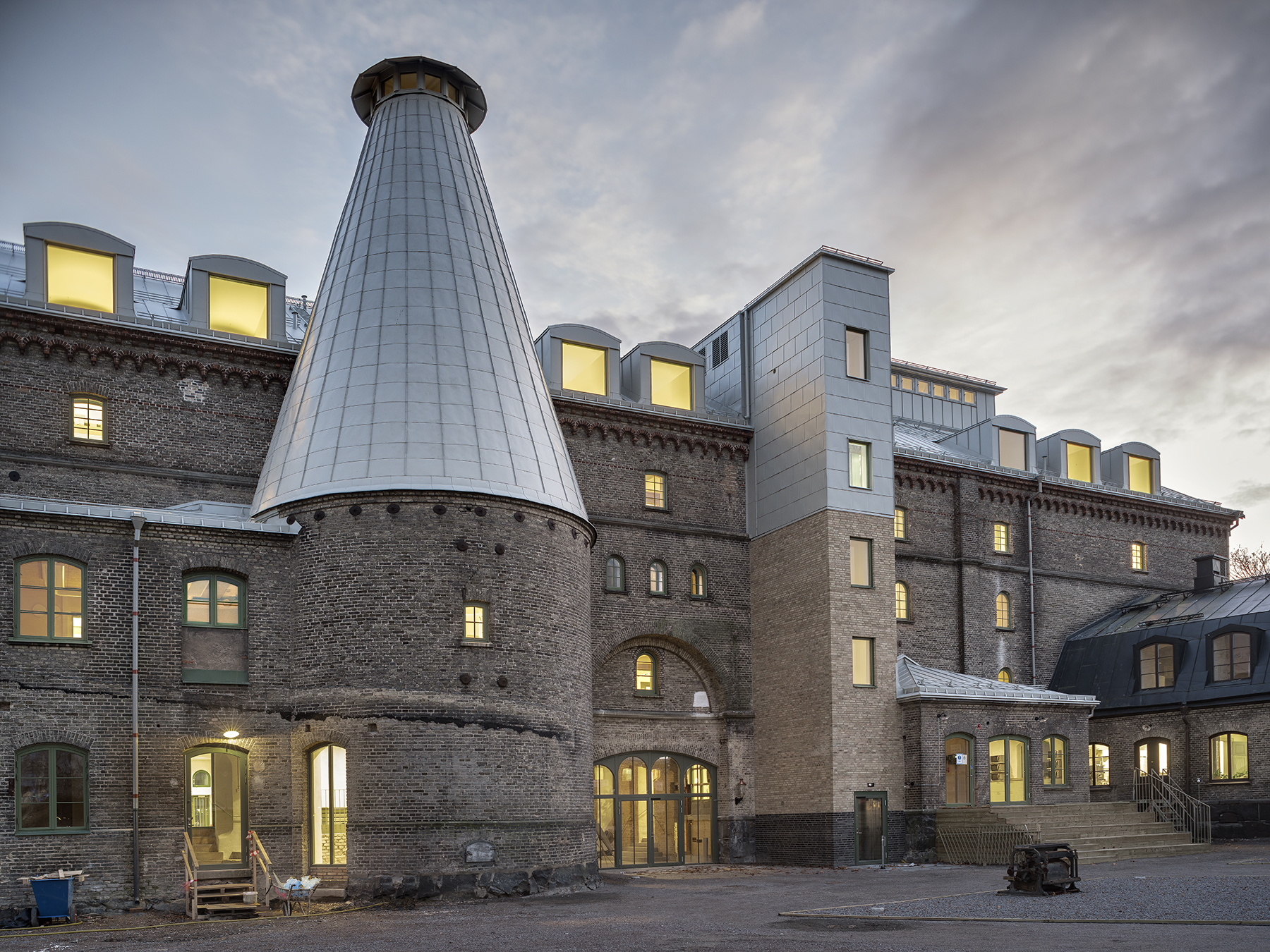 En äldre tegelbyggnad med ett stort koniskt torn och modernt tillägg på taket, fotograferad i skymningen med ljus i fönstren.