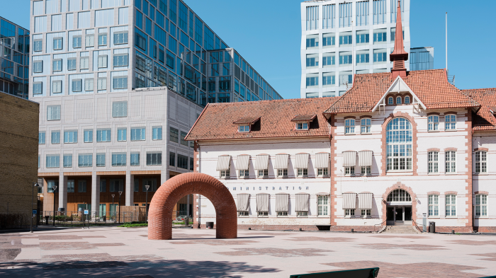 En äldre administrationsbyggnad med rött tegeltak står bredvid moderna kontorshus av glas, med en båge i tegel framför.