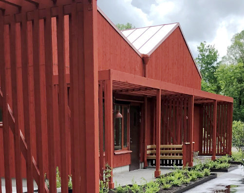 En röd träbyggnad med ett pergolatak, en bänk vid ingången och träd i bakgrunden under en molnig himmel.
