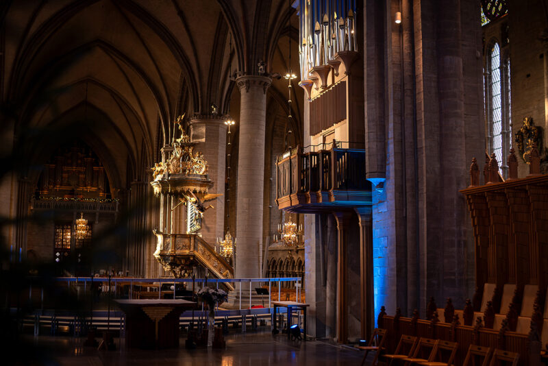 Interiör av en stor katedral med valvade tak, predikstol, orgel, ljuskronor och rader av trästolar under belysning.