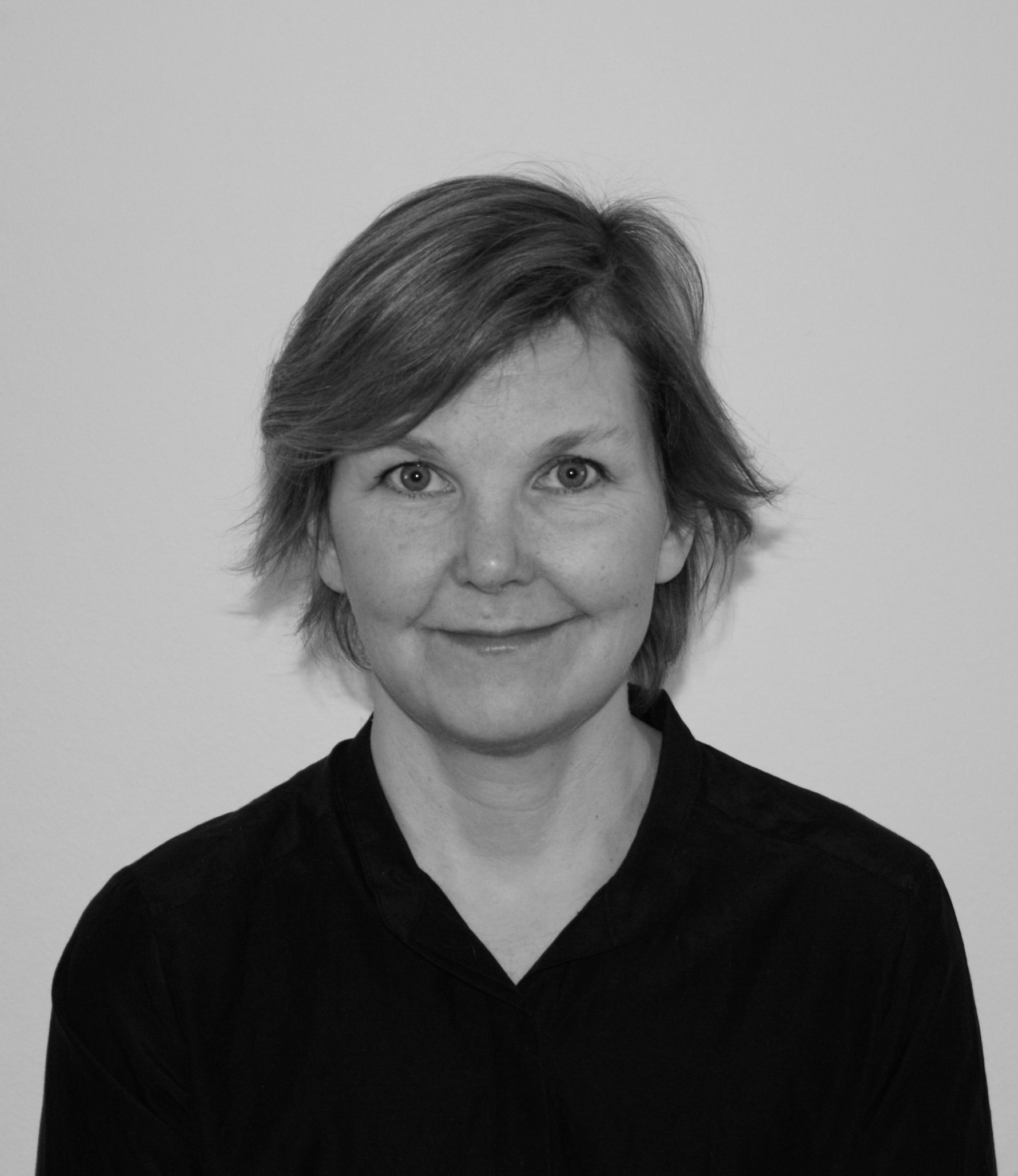 Porträtt i svartvitt av en kvinna med kort hår och mörk tröja, som tittar mot kameran och ler svagt mot en enkel bakgrund.