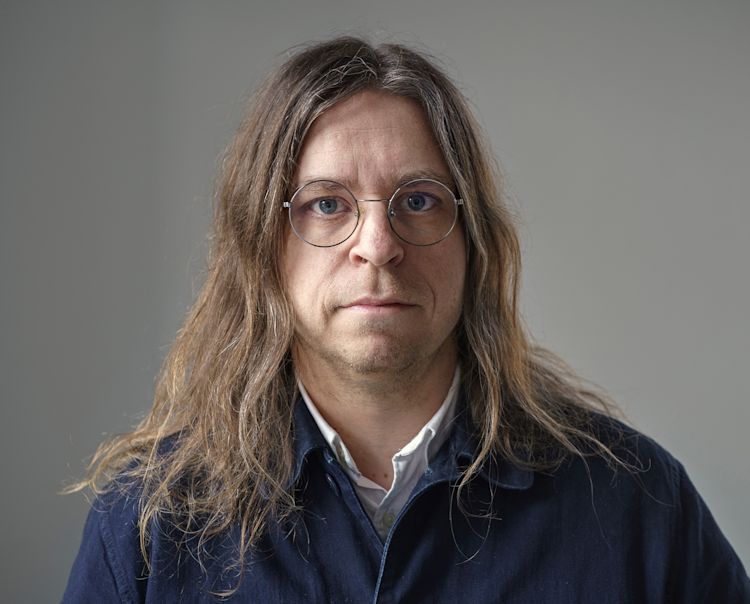 En person med långt brunt hår och glasögon, iförd mörkblå jacka över vit skjorta, står framför grå bakgrund.