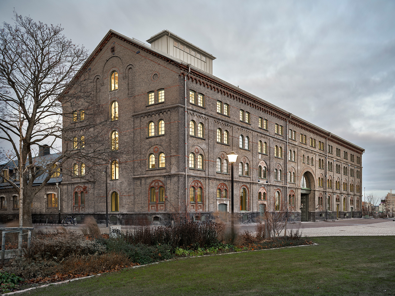 Fyra våningar högt tegelhus med bågformade fönster, fönstren lyser. Nakna träd och gräsyta runt huset under molnig himmel.