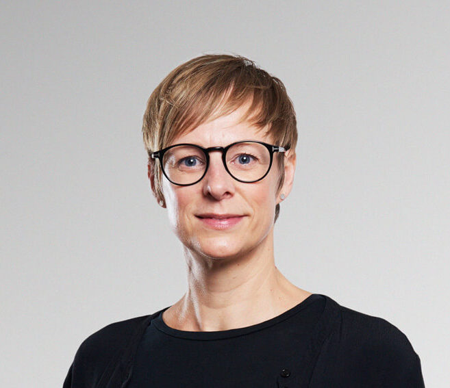 En person med kort blont hår, runda svarta glasögon och svart topp står framför en ljusgrå bakgrund med neutral min.