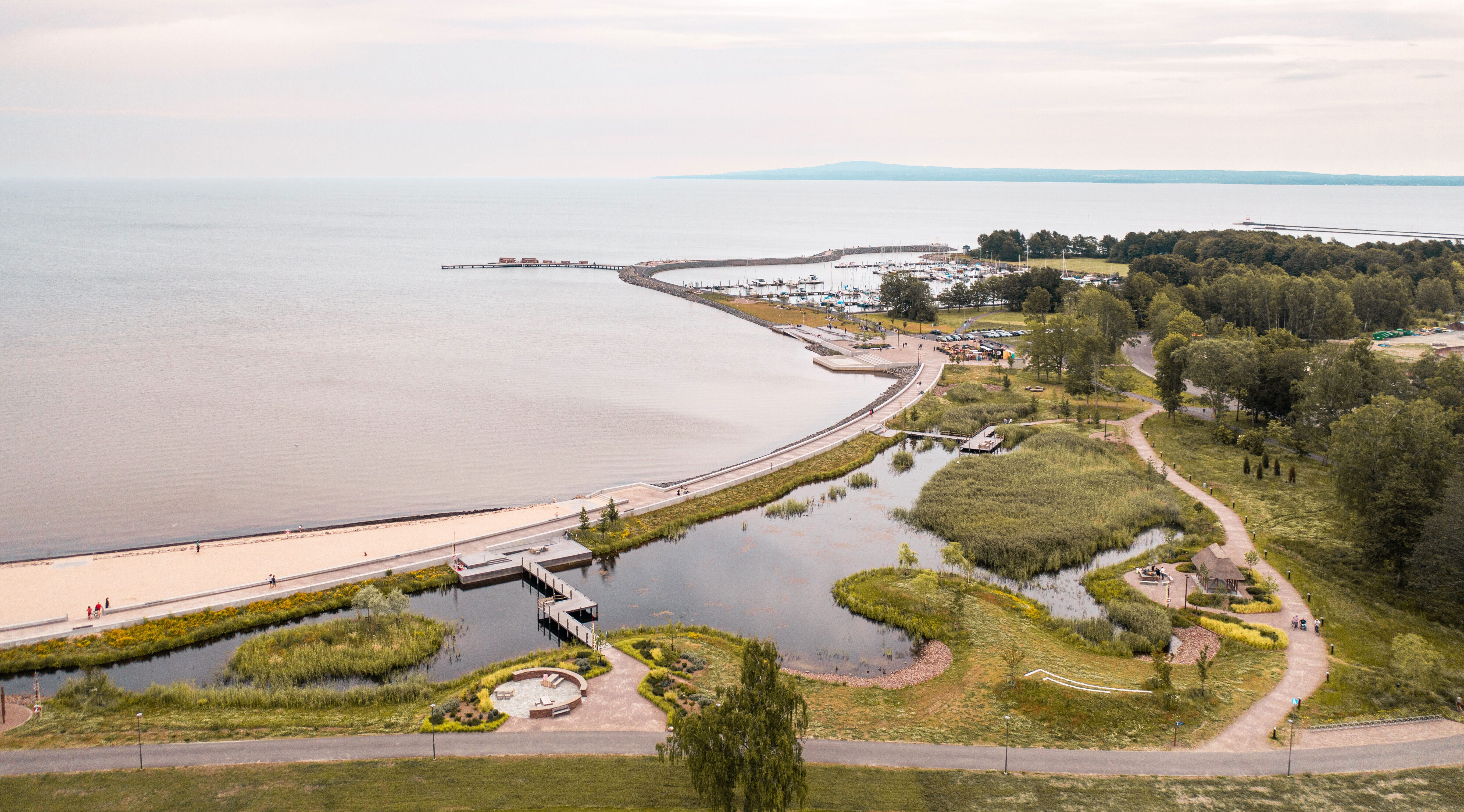 Flygbild av en kust med sandstrand, gångvägar, grönska, en liten damm och en böjd brygga som går ut i vattnet under molnig himmel.