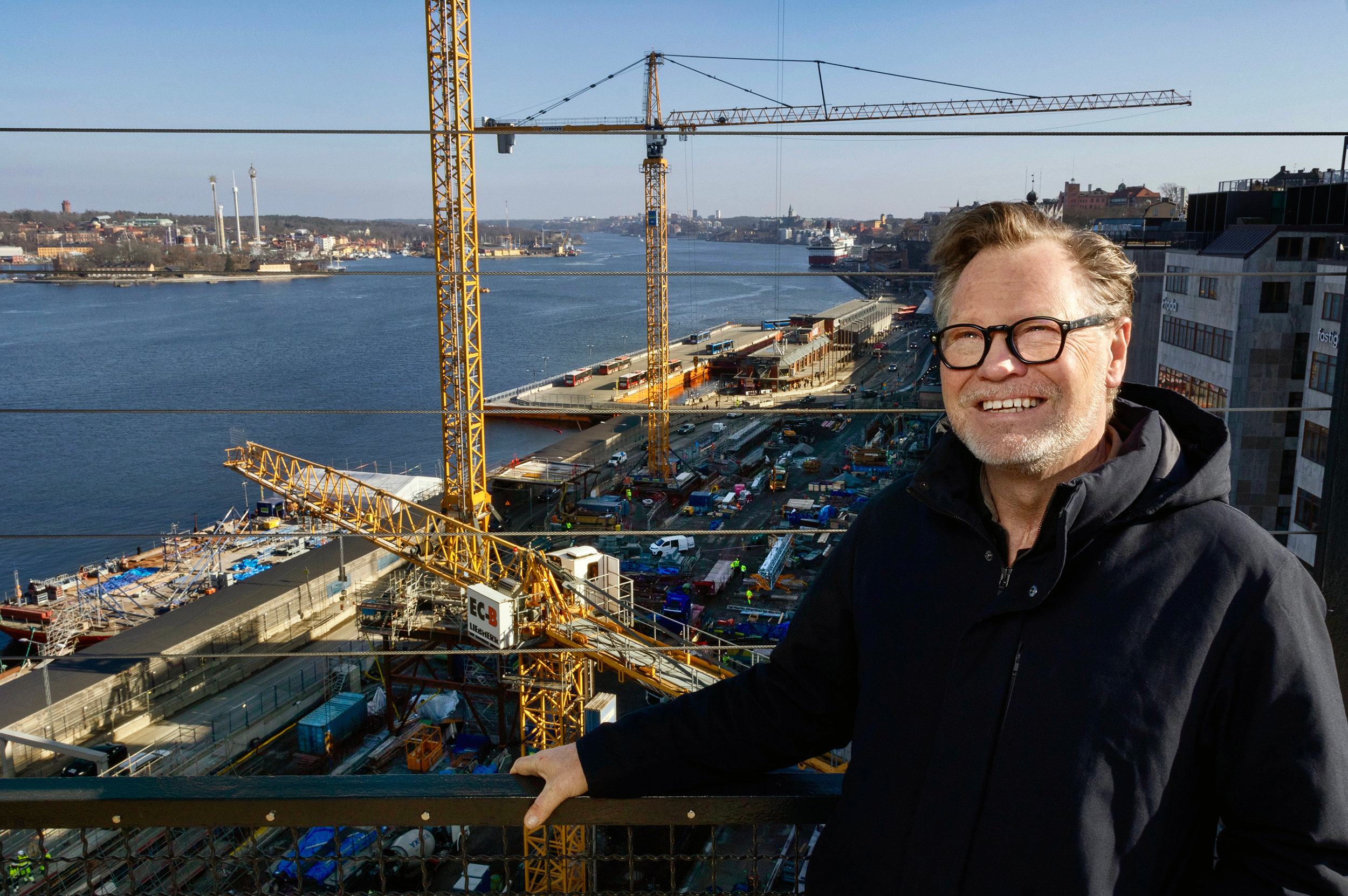 En man med glasögon och mörk rock står på en balkong som vetter mot en flod och ett stort byggområde med kranar.