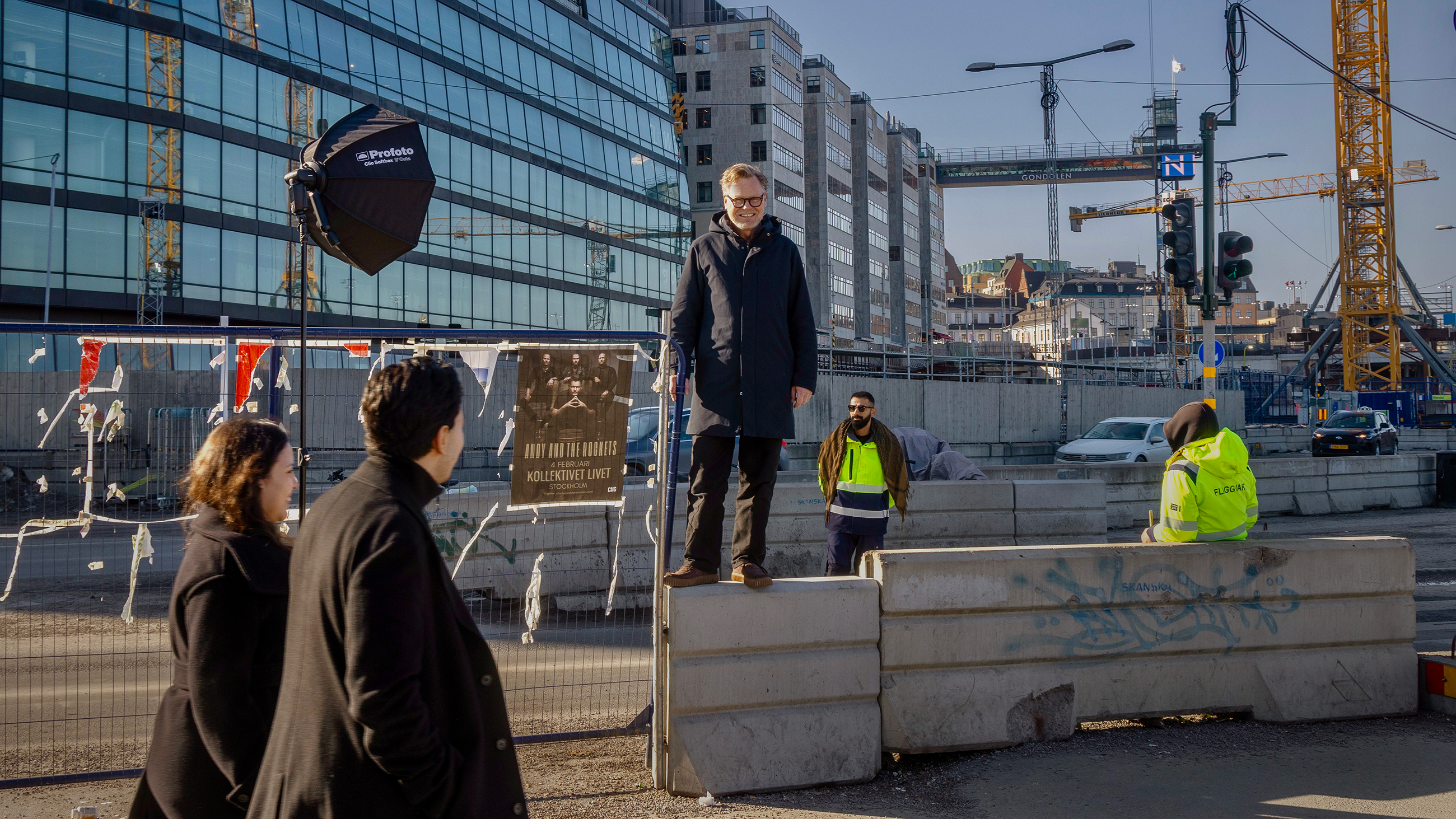En man i rock står på ett betongblock vid en byggarbetsplats och talar till tre personer. Två arbetare i reflexvästar. Byggnader och kranar syns.