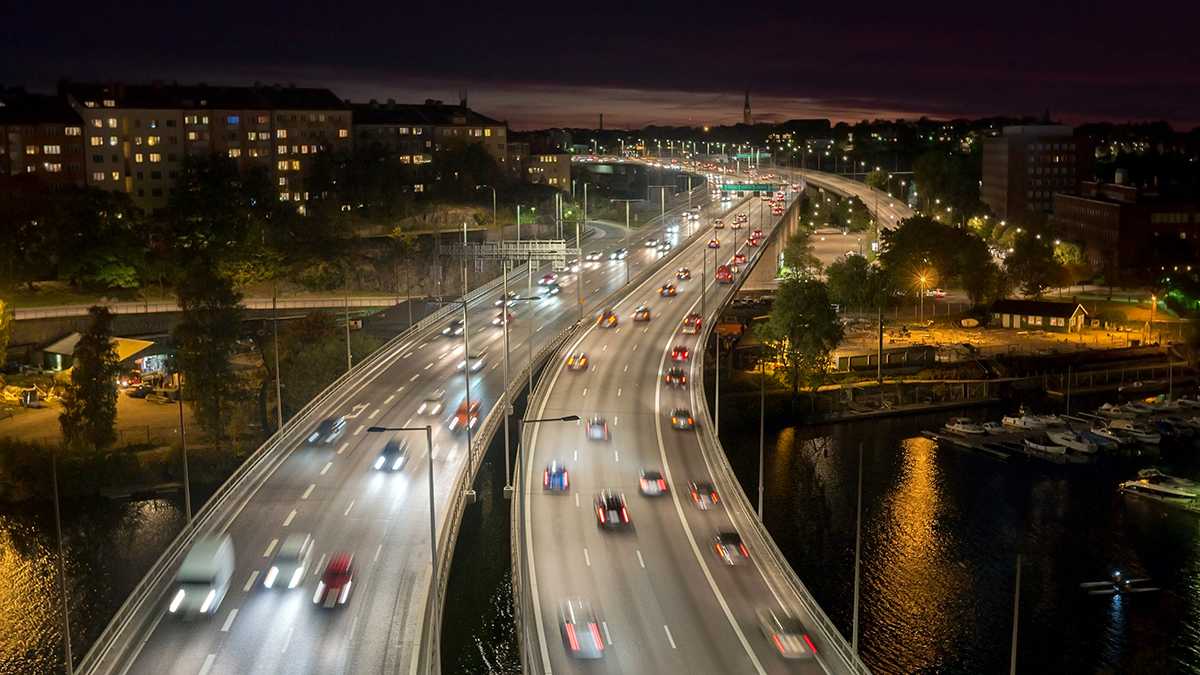 Flygfoto av en flerfilig motorväg på natten med bilar i rörelse, stadens byggnader och gatlyktor synliga i bakgrunden.