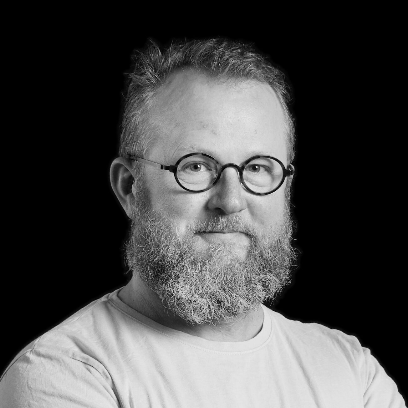 En man med skägg, glasögon och kort hår har på sig en ljus skjorta och står mot en svart bakgrund.