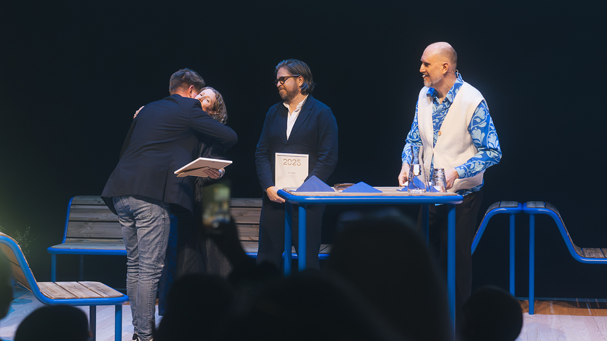 Fyra personer står på scenen; en person kramar en annan och håller ett certifikat, två tittar på, en står vid ett bord med böcker och papper.