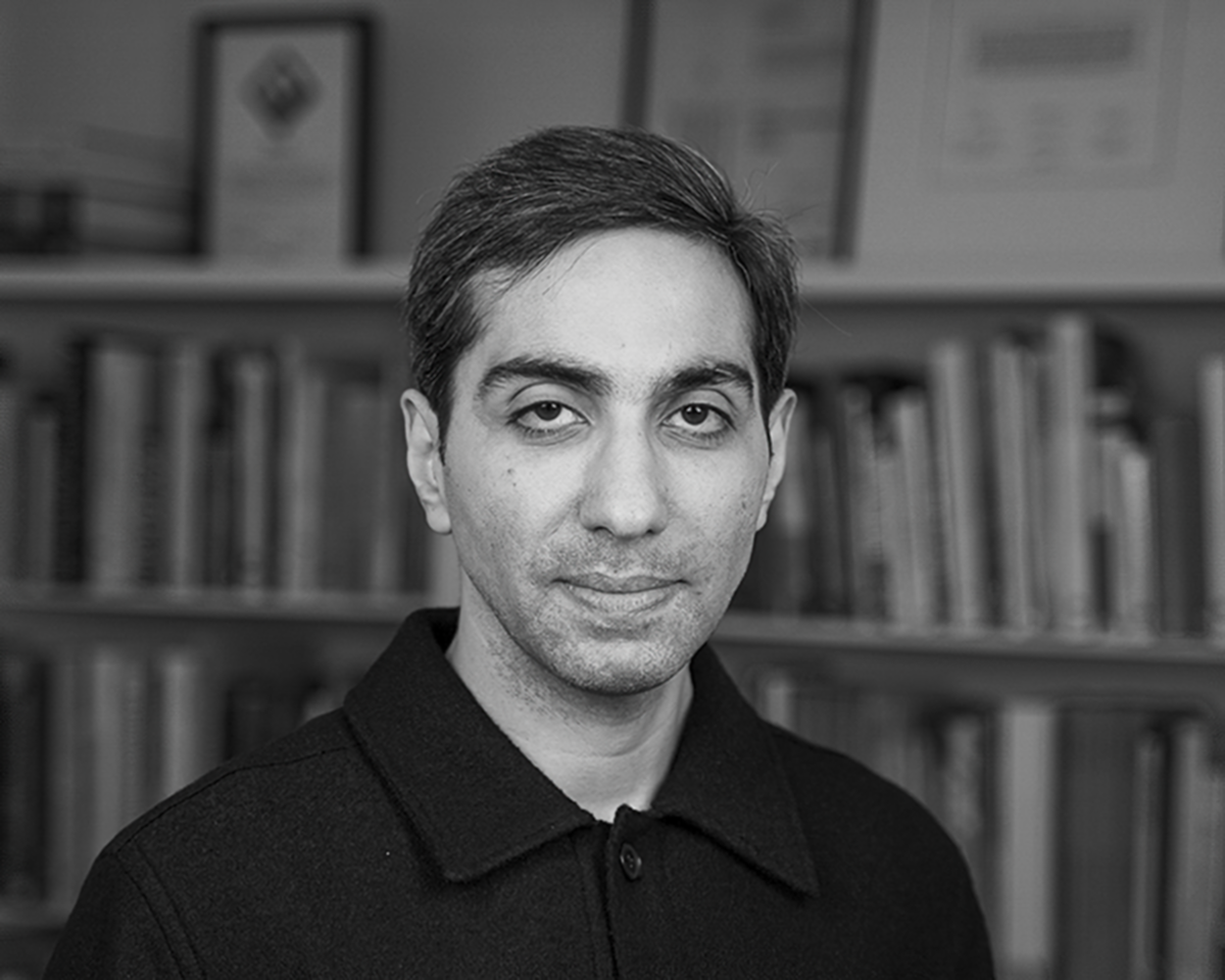 En man med kort mörkt hår och skjorta med krage står framför en bokhylla med böcker och inramade dokument.