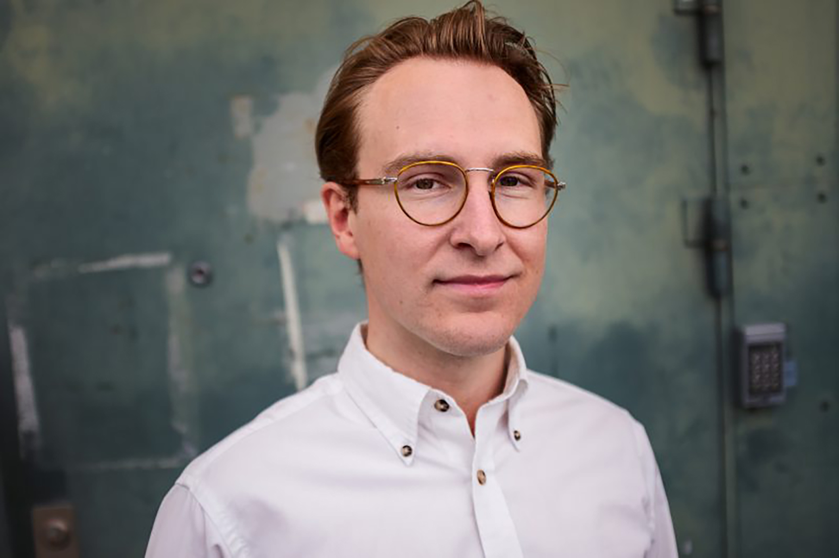 En man med ljusbrunt hår och glasögon har en vit skjorta och står framför en vägg som ser ut att vara industriell.