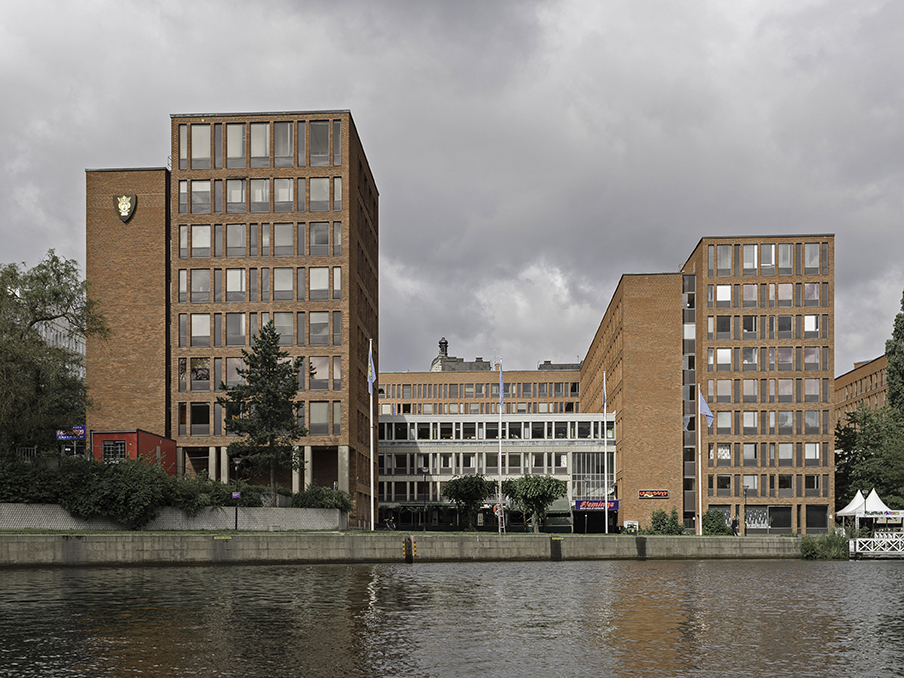 Kontorsbyggnader av rött tegel med stora fönster står vid en flod. Framför huvudentrén finns ett träd och flaggor.